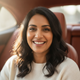 Headshot of a smiling South Asian woman in her 30s, in a car, looking at the camera.